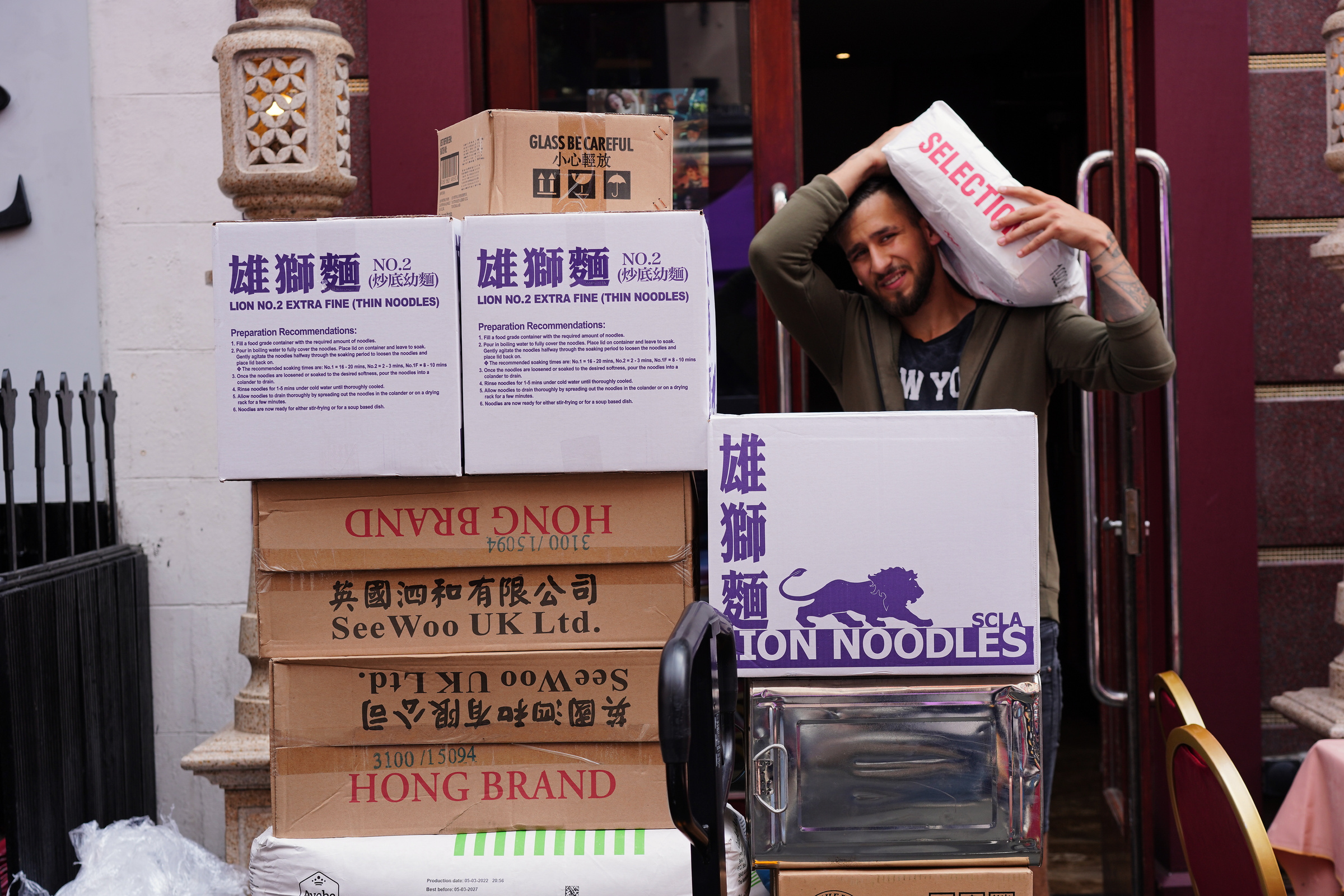A man carries a white sack beside a stack of cardboard boxes.