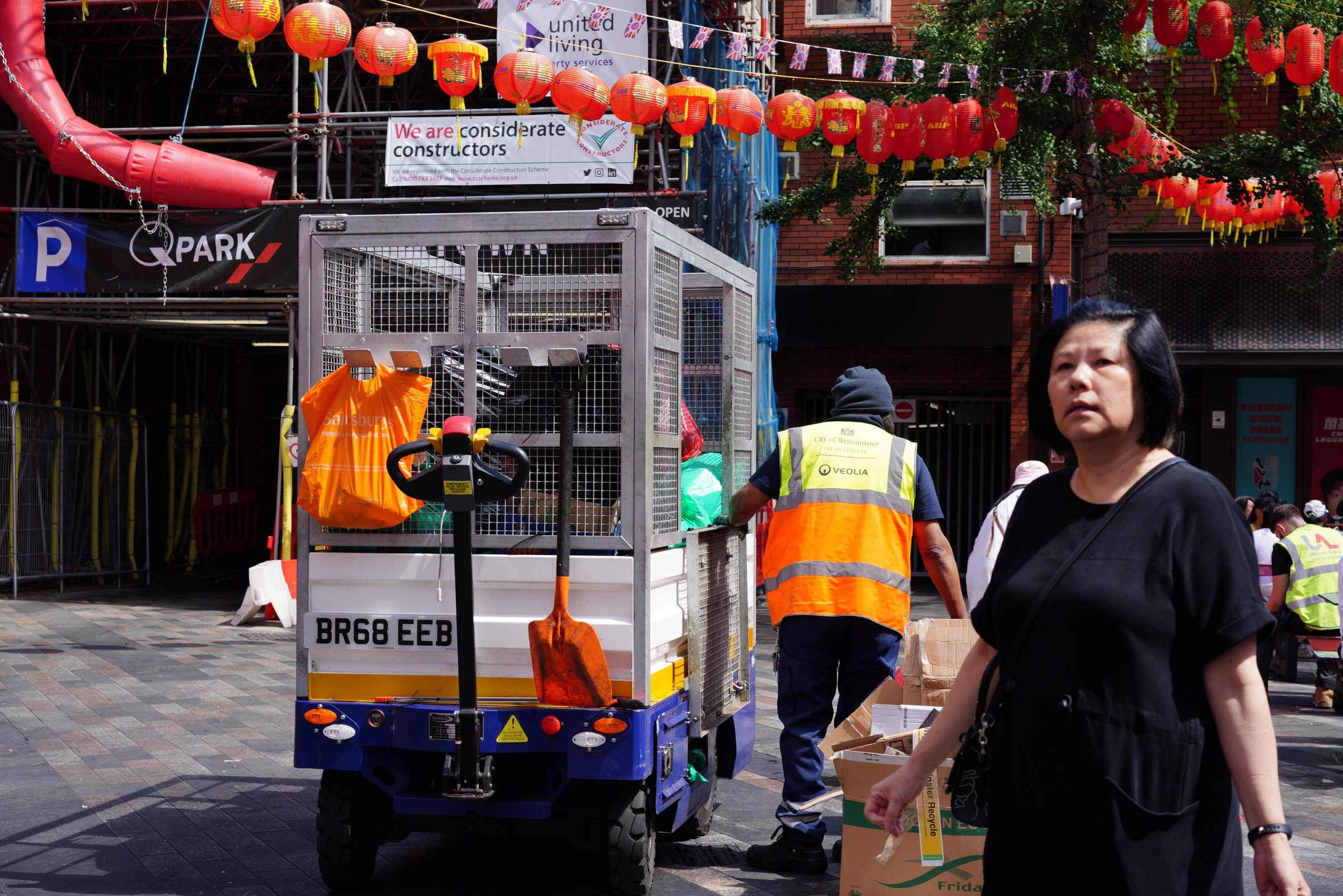 A cleaning vehicle, passing figures and construction equipment occupy the street beneath lanterns.