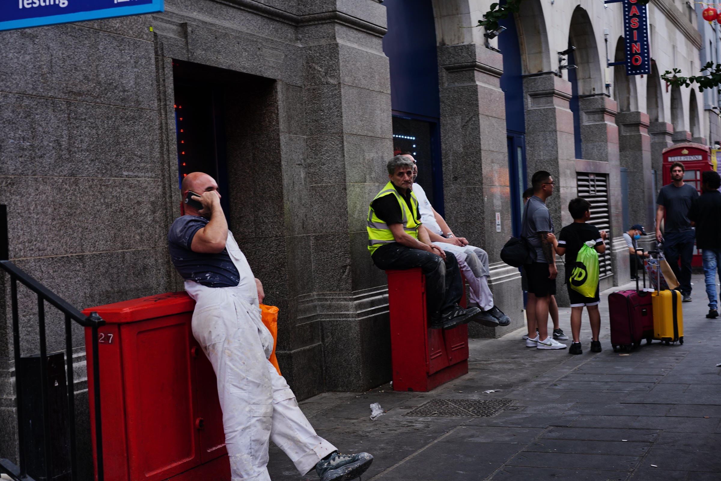 People sit and lean on red street cabinets beside a stone building.