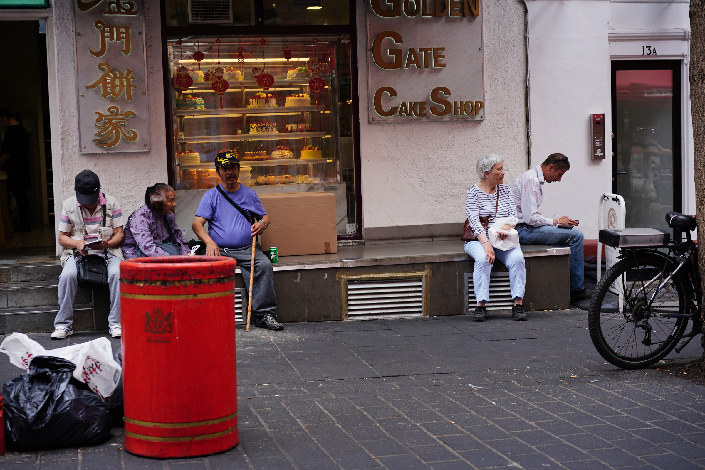 Five people sit outside a cake shop behind a red street bin.