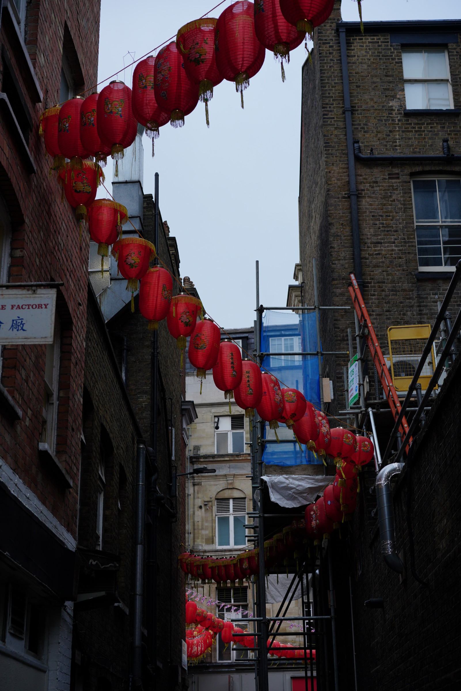 Strings of red lanterns cross a narrow opening between brick walls, pipes and scaffolding.