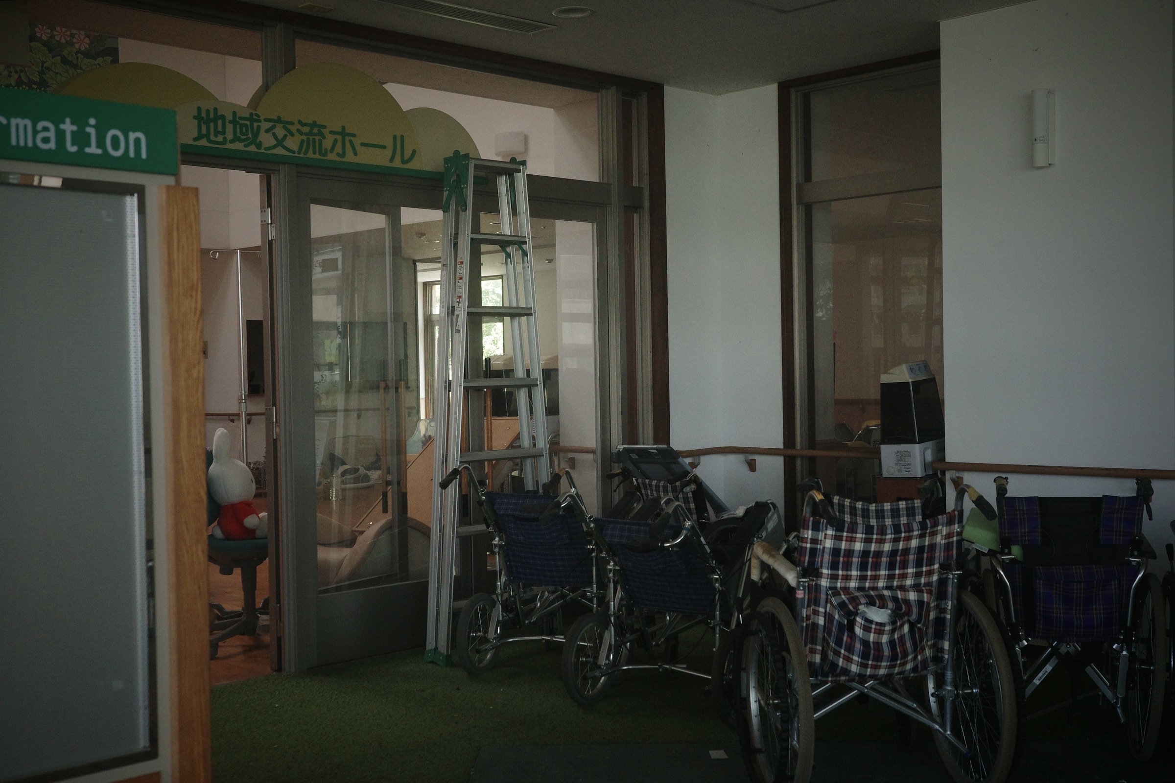 Wheelchairs gather beside glass doors, a ladder and a toy rabbit in a deserted interior.