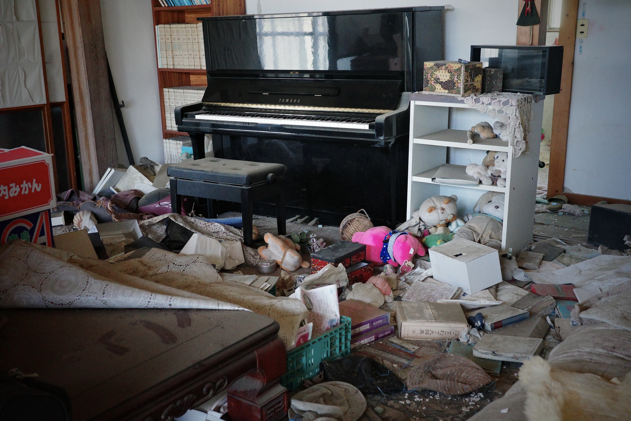 An upright piano and a shelf of soft toys stand among scattered papers and household objects.