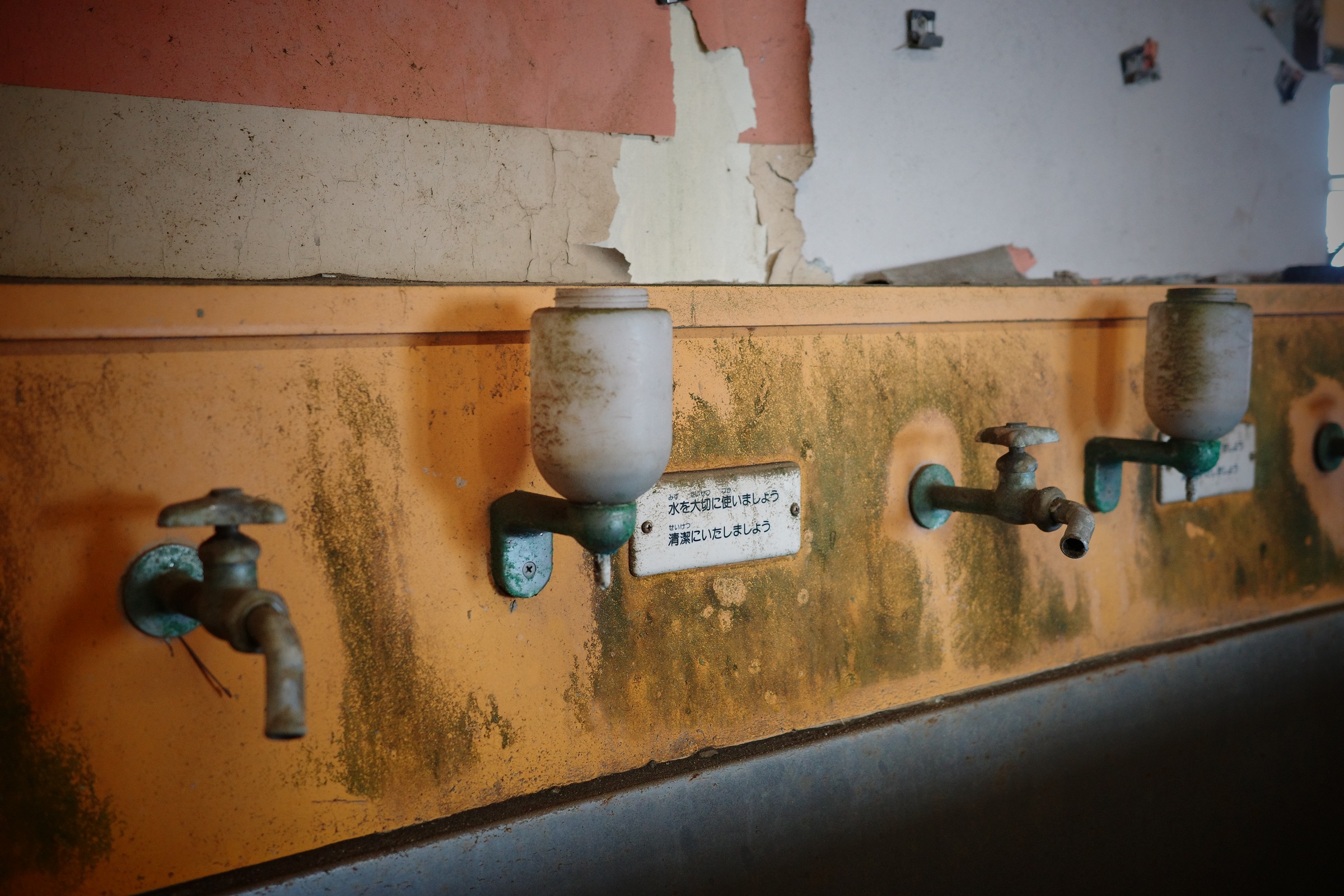 A row of taps and soap containers lines a wall with peeling pink and ochre paint.