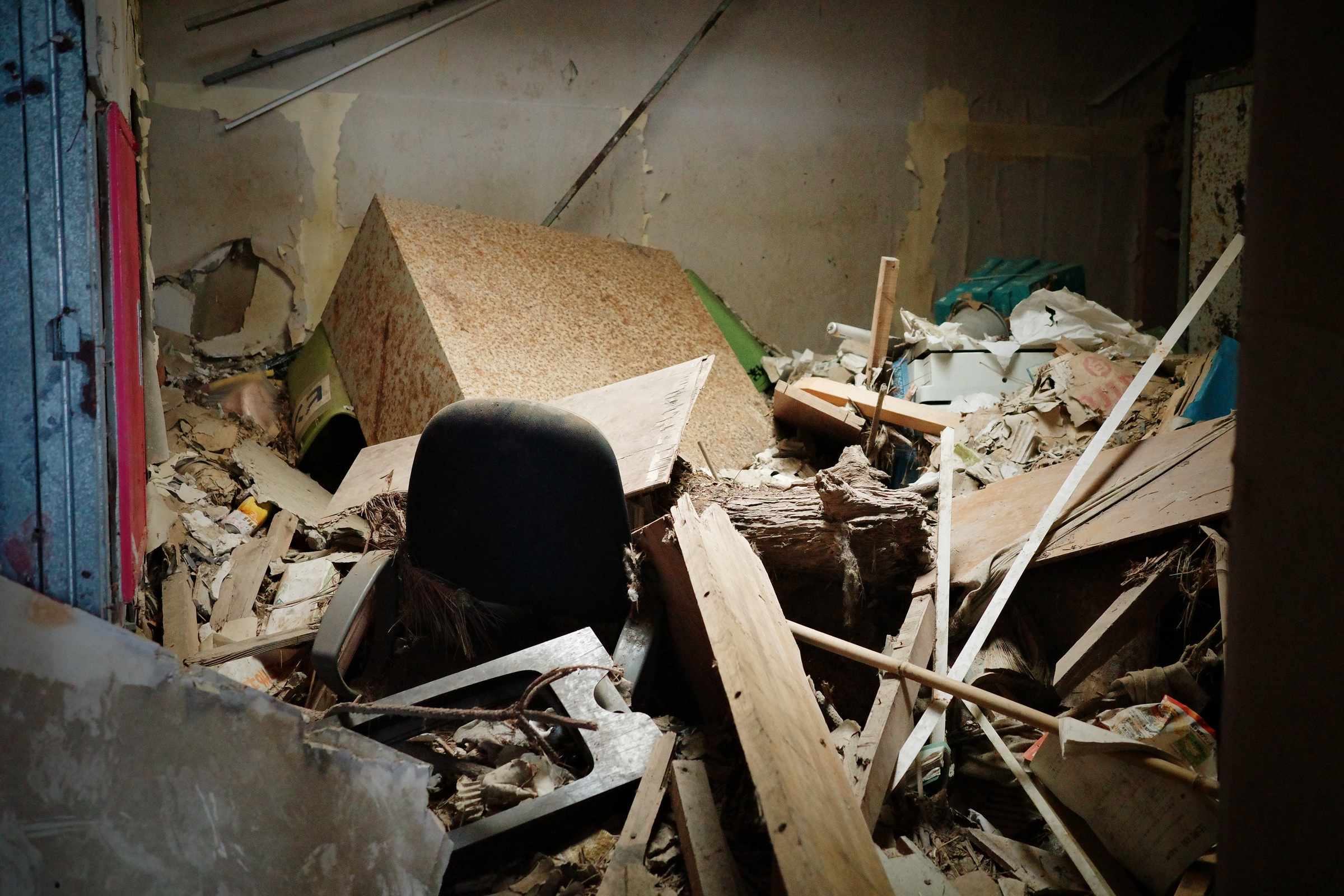A black chair is surrounded by fallen boards and broken plaster, framed by a blue doorway.