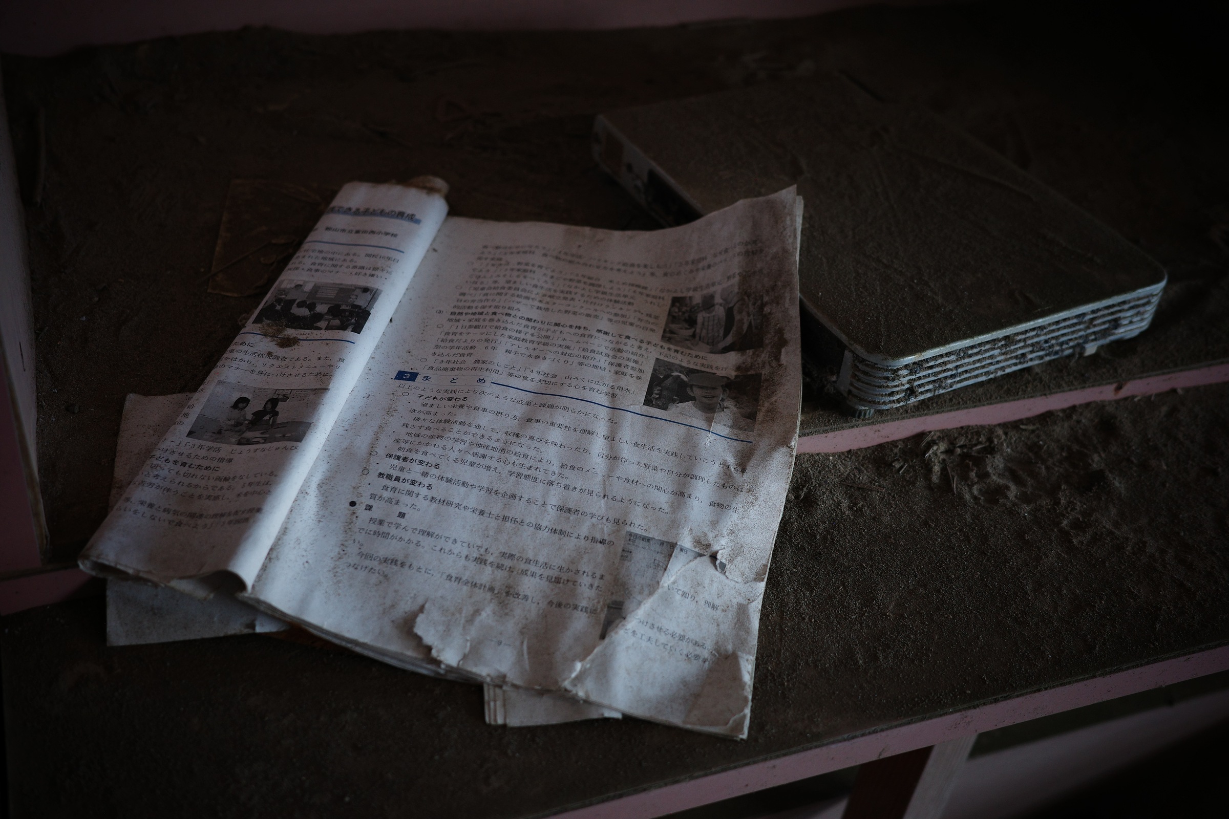 An open illustrated Japanese document rests on a dusty shelf beside stacked laptops.