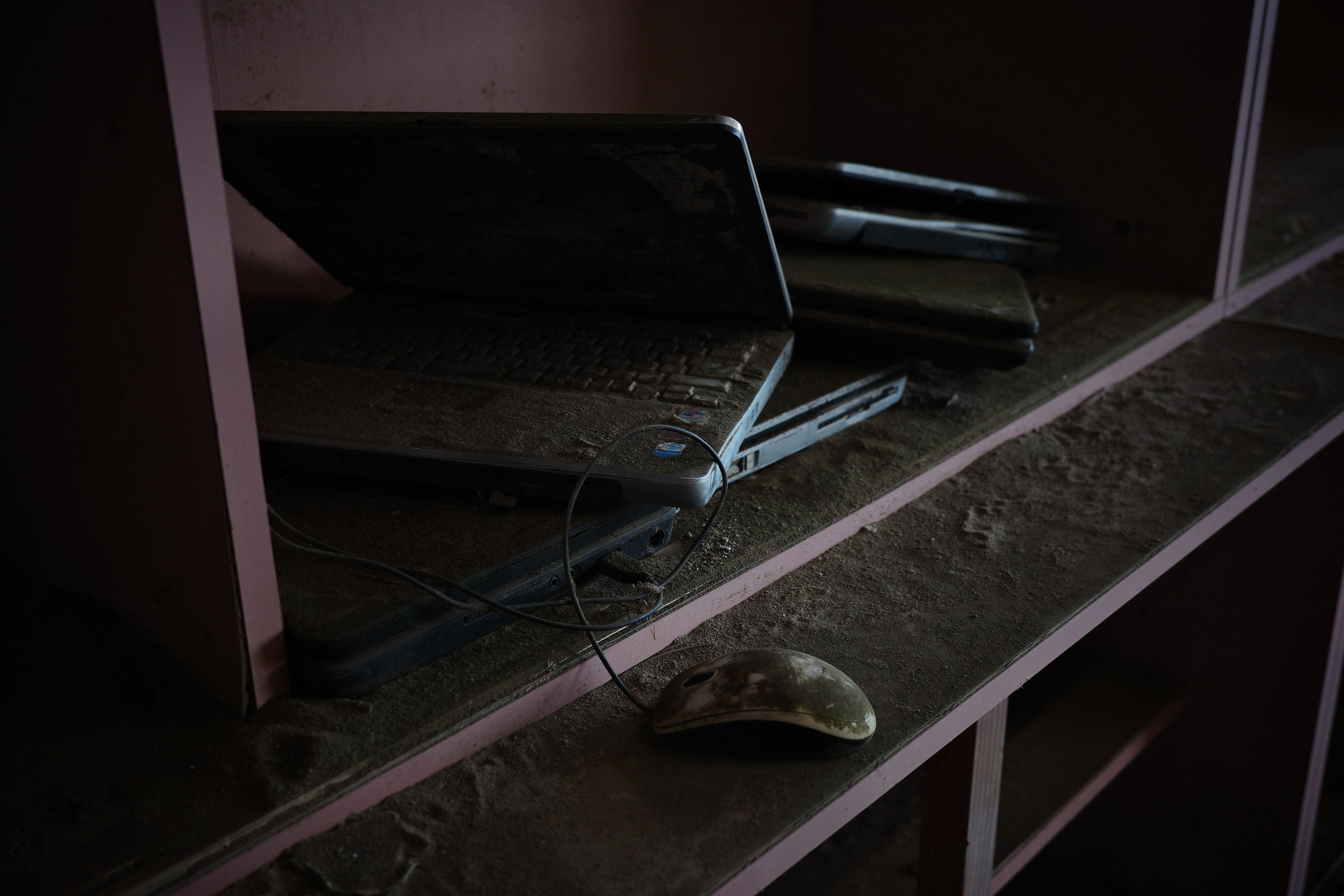 An open laptop, stacked computers and a wired mouse lie on dust-covered shelves.