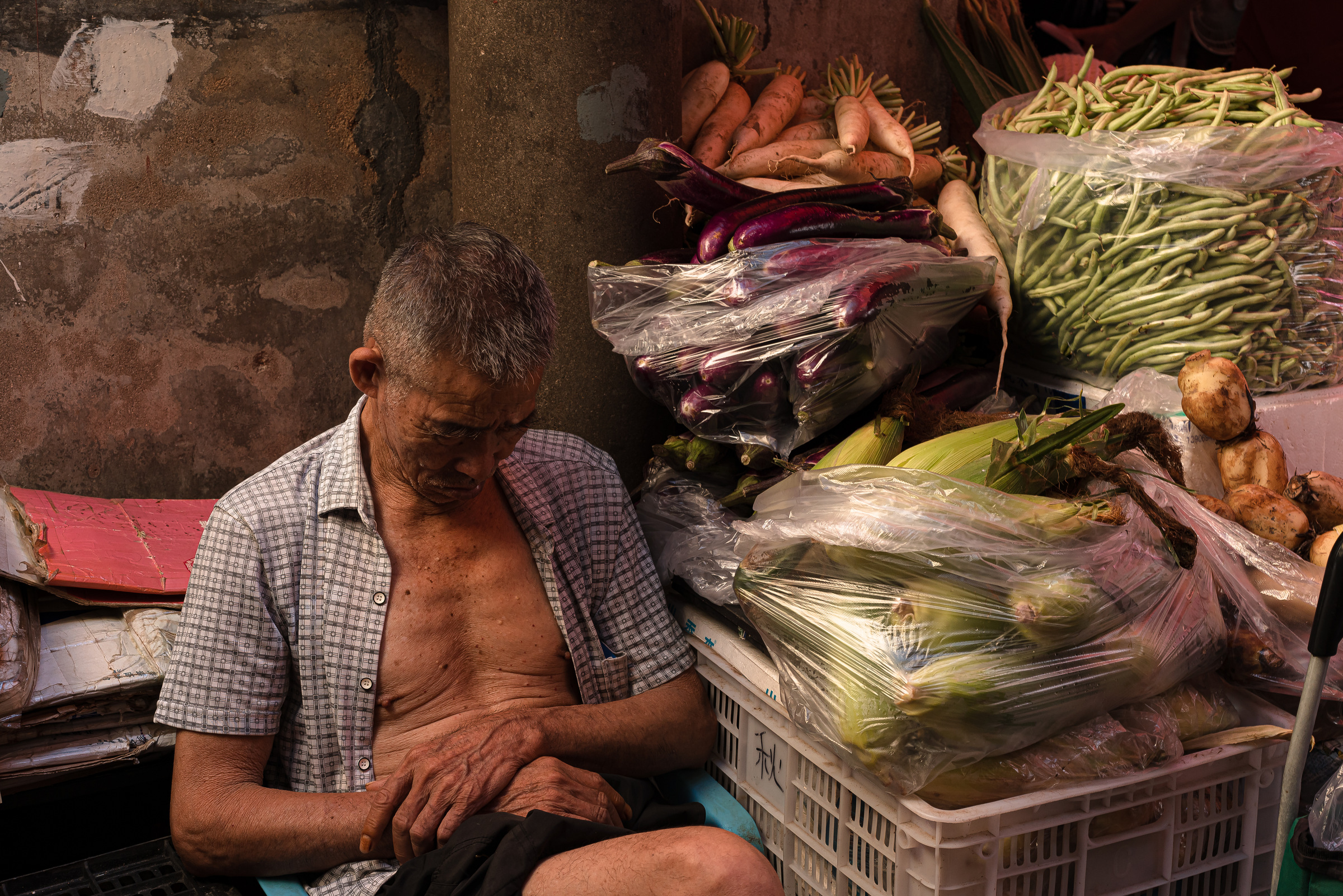 A seated market vendor rests with his head lowered beside bags of vegetables.