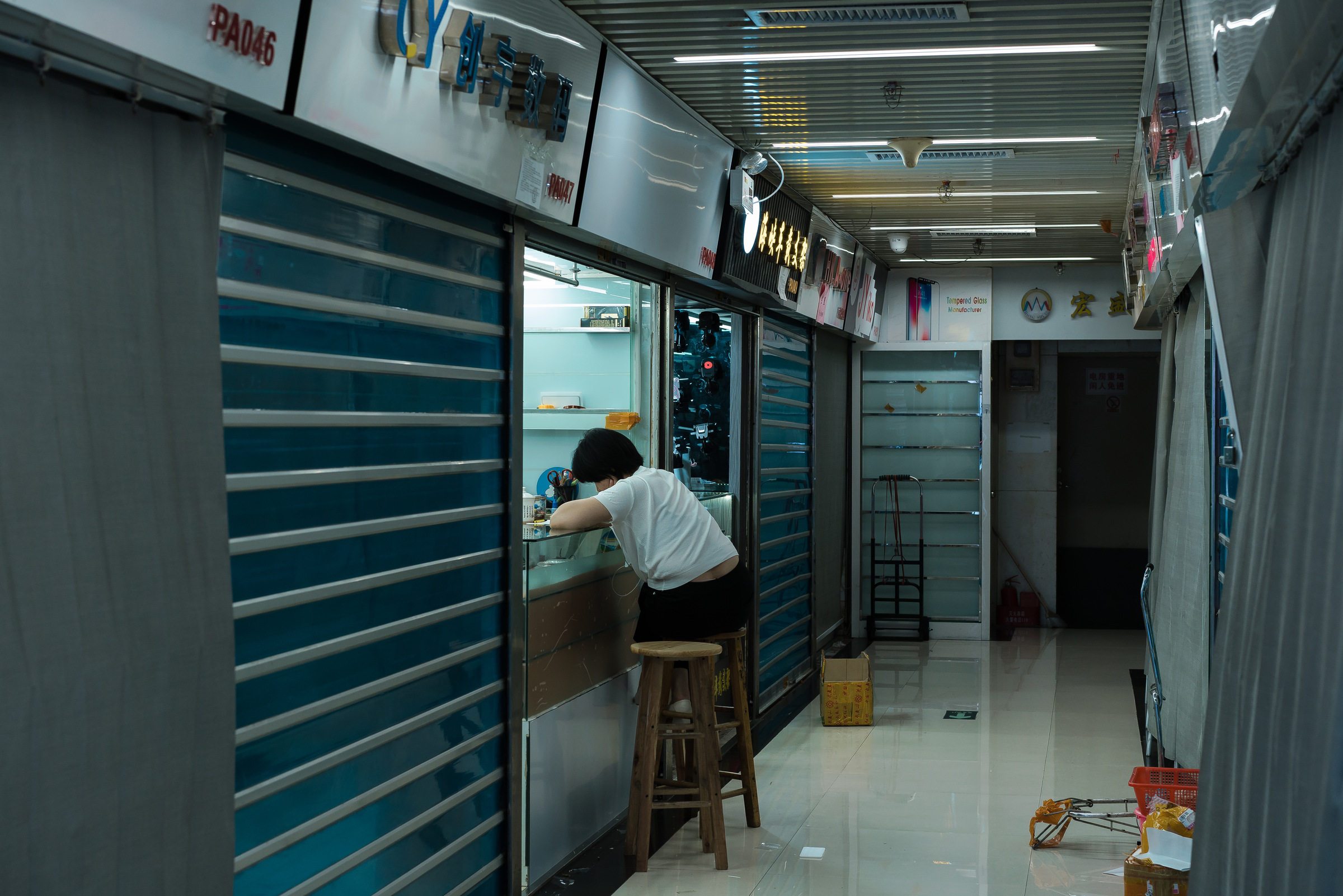 A person leans over a counter beside blue shutters in a commercial arcade.
