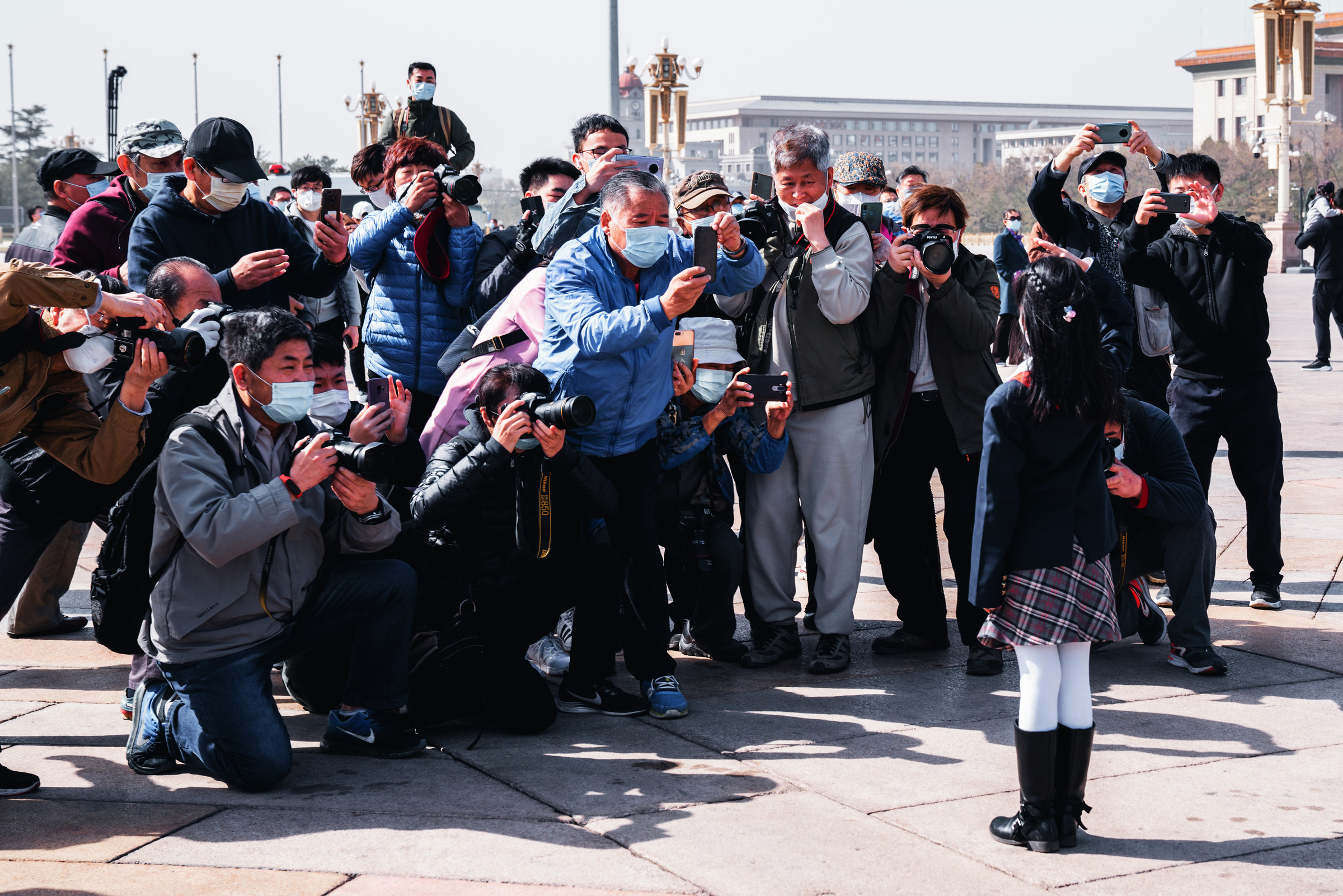 Masked photographers kneel and raise their cameras toward a girl in a public square.