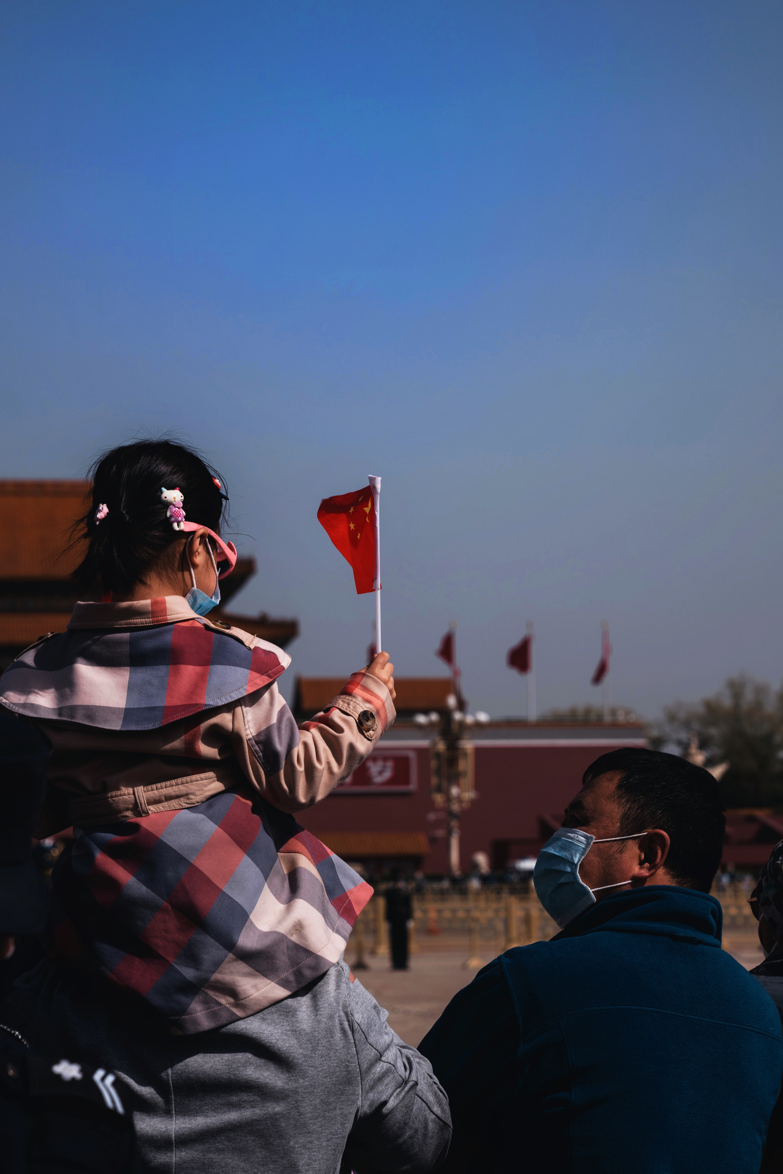 A girl sits on an adult’s shoulders holding a small Chinese flag near Tiananmen.