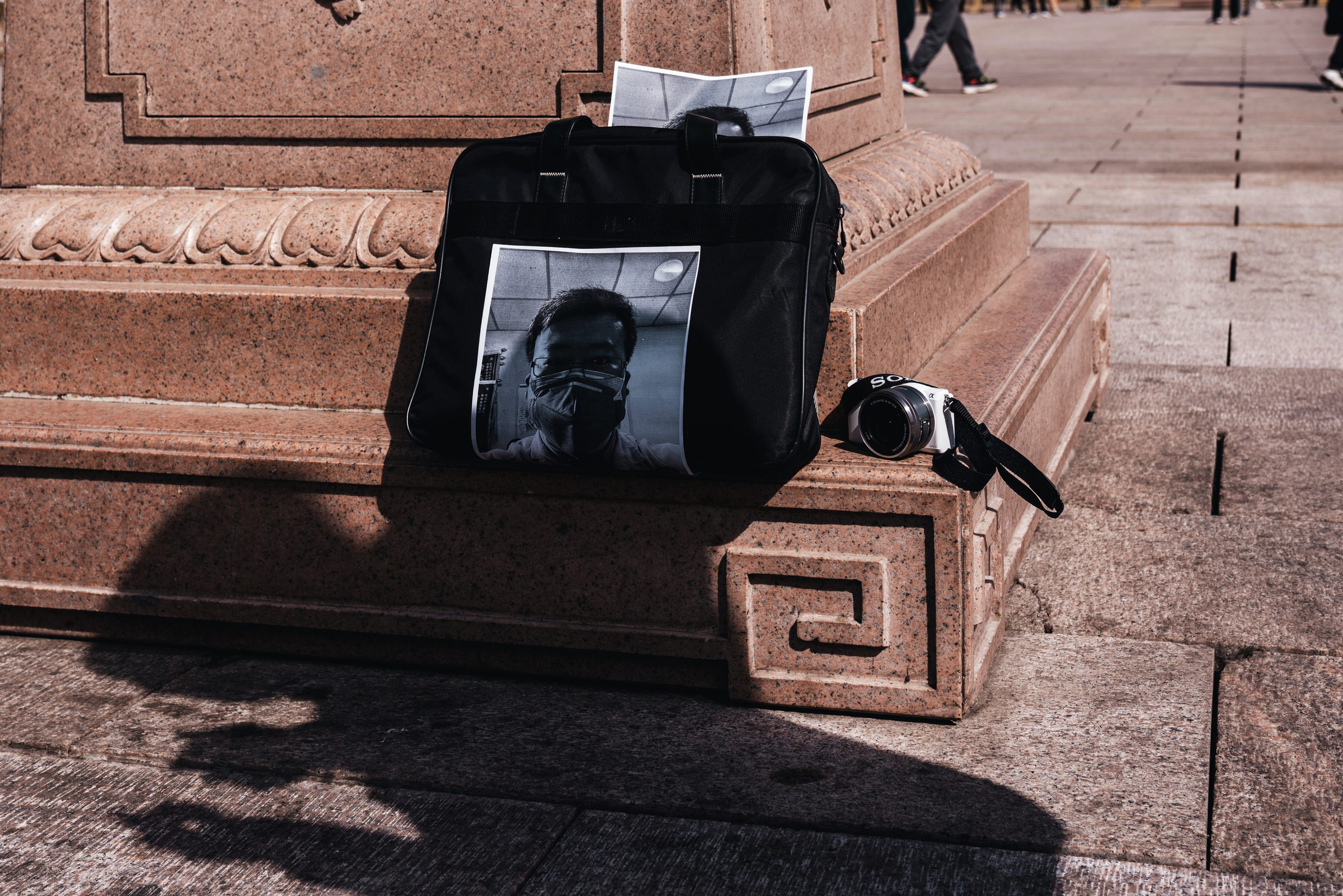 Printed photographs of Li Wenliang rest against a black bag on a stone plinth, with a camera beside them and a person’s shadow below.