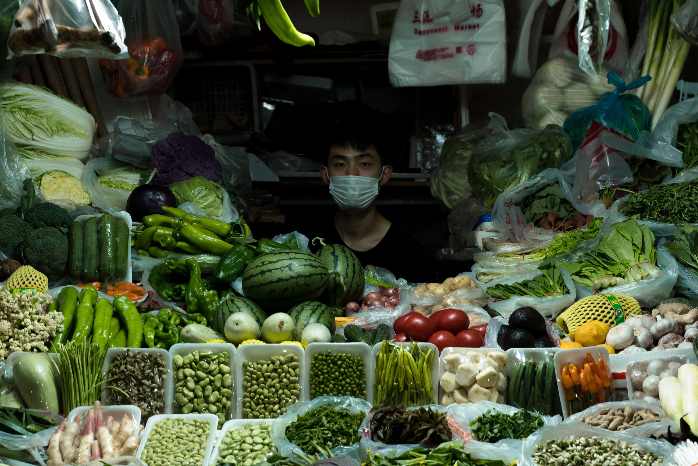A masked vendor looks outward from behind densely arranged vegetables.