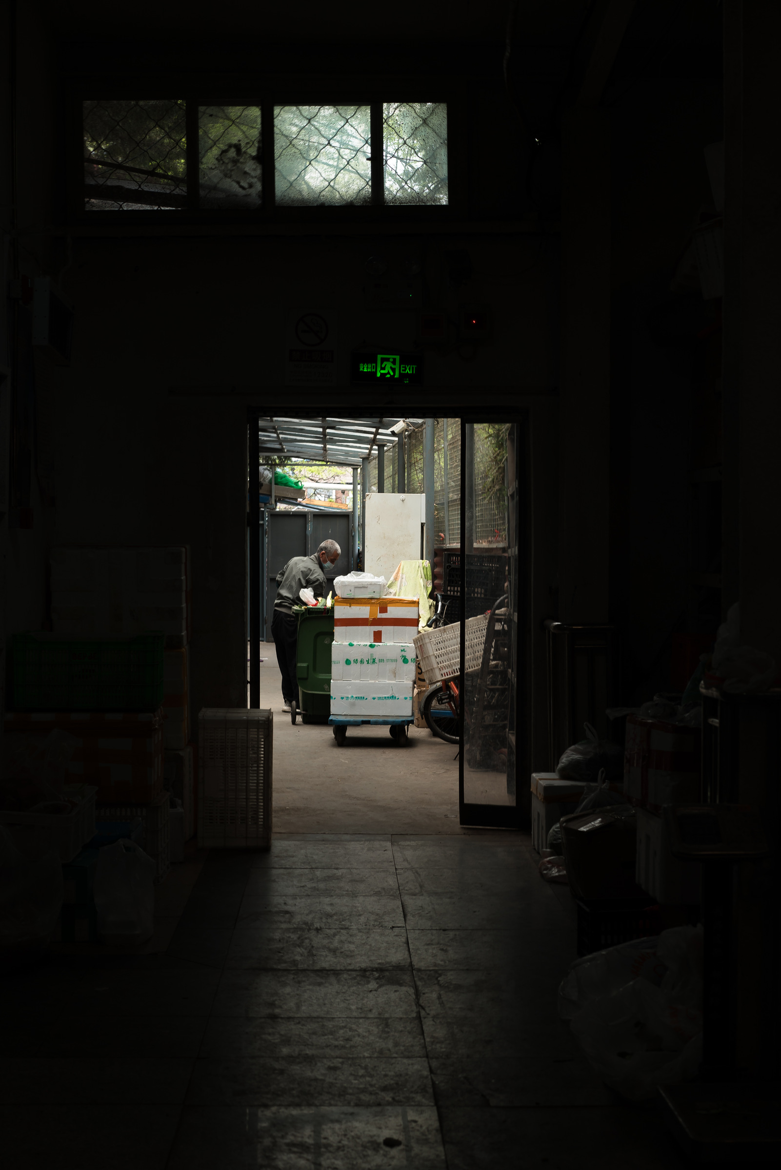 A dark market passage opens toward a person, boxes and a cart beyond a doorway.