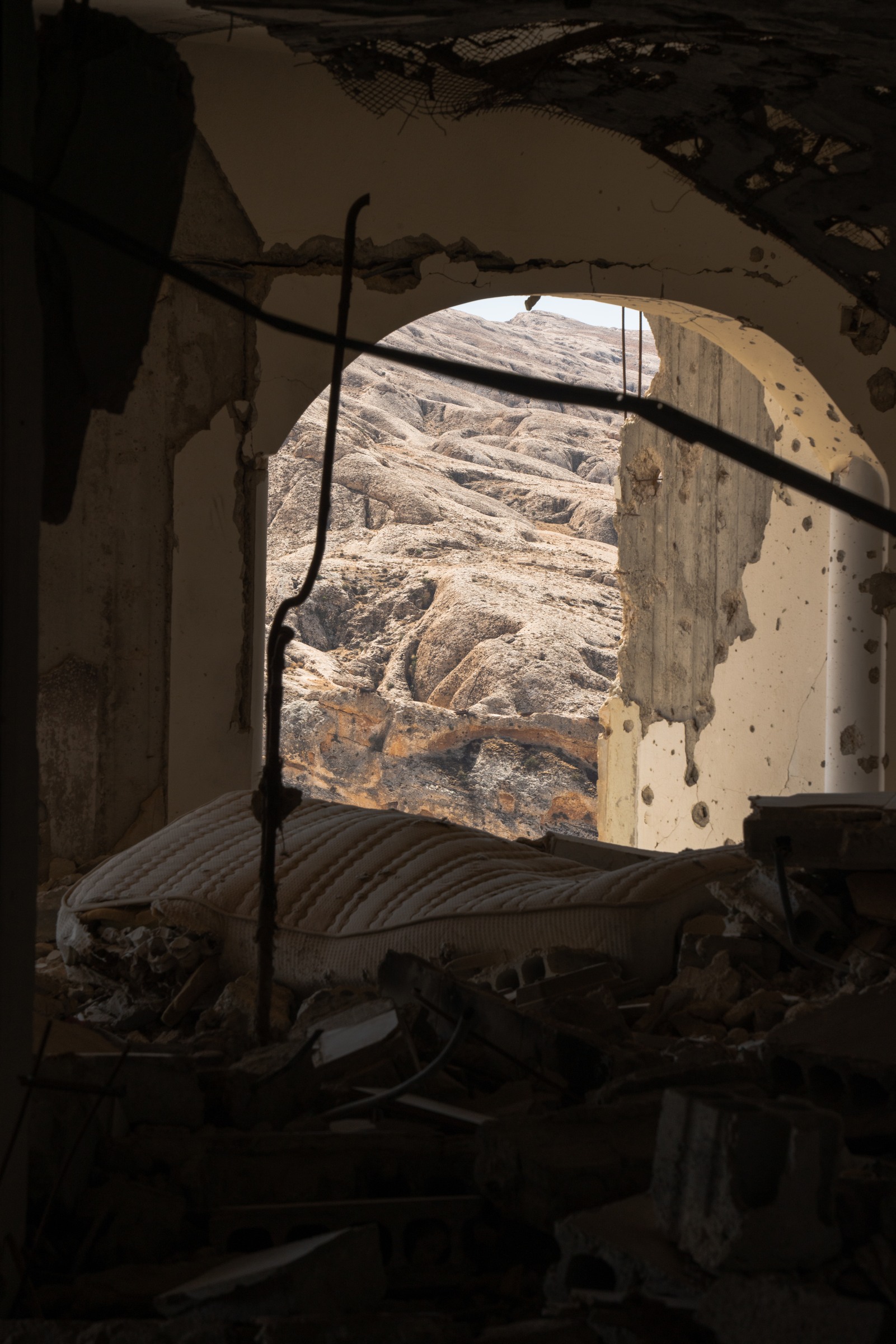A mattress lies beneath a broken arch opening onto sunlit rocky hills.