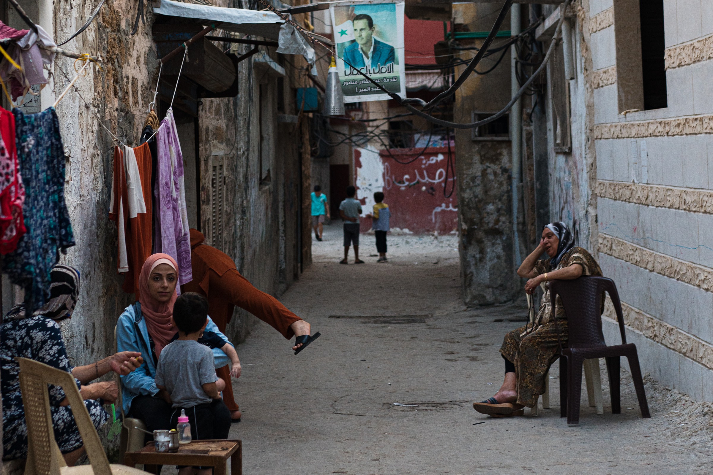Women and children occupy a narrow alley beneath hanging fabrics, cables and a portrait.