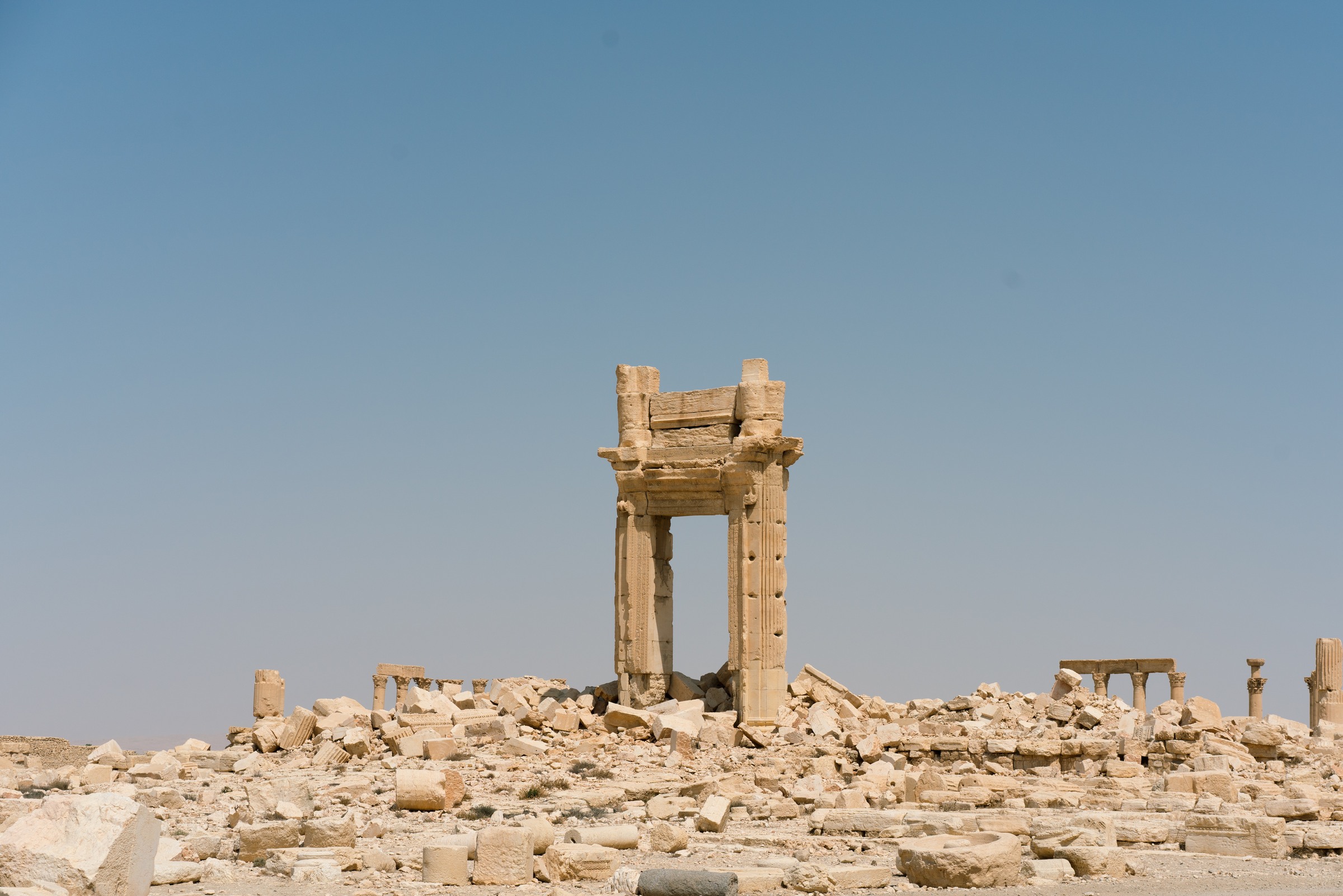 A stone gateway stands among fallen architectural fragments beneath a wide blue sky.