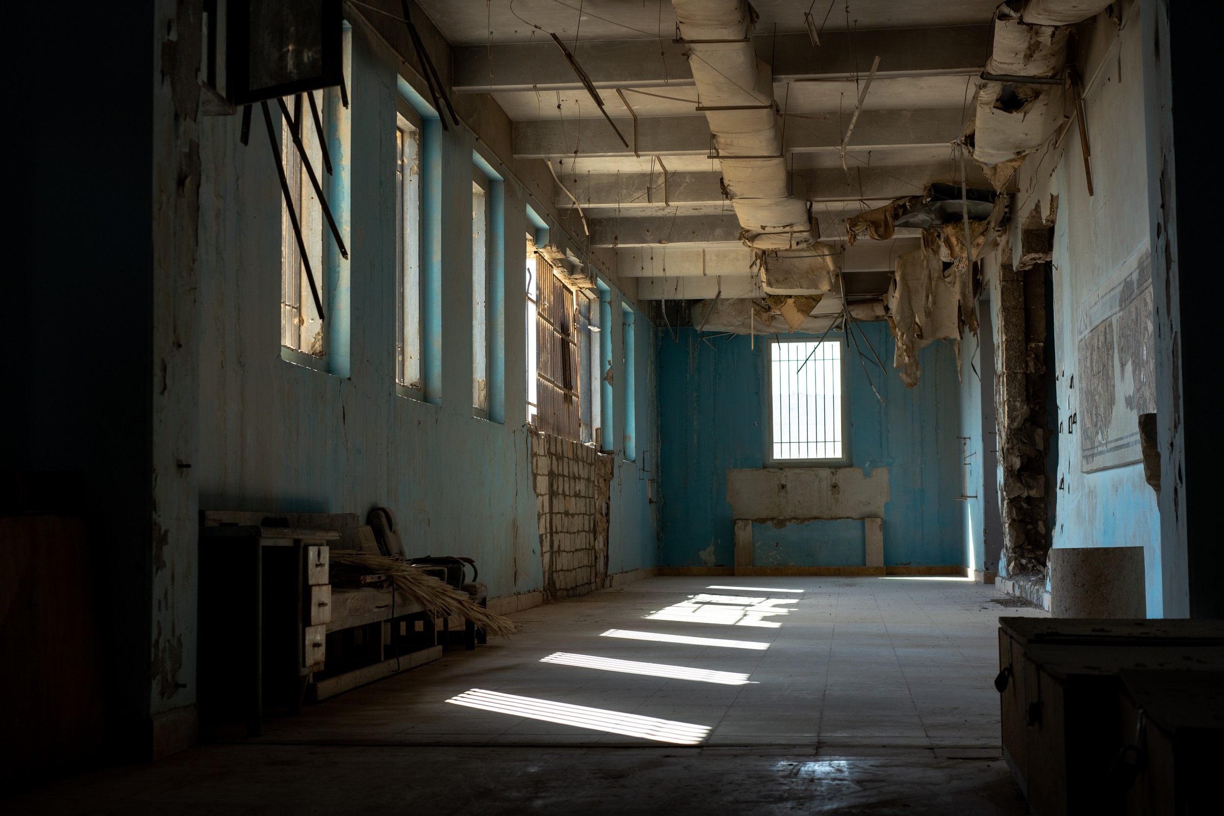 Window light crosses the floor of a blue gallery beneath damaged ceiling ducts.